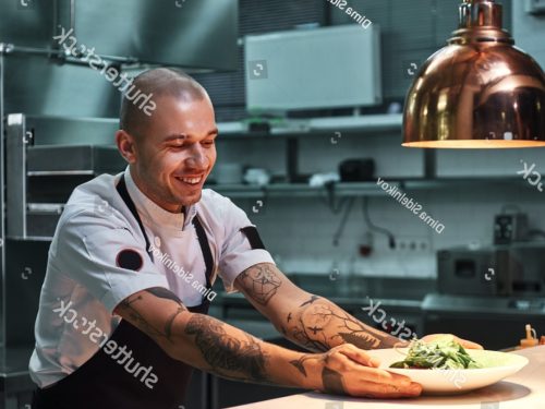 stock-photo-ready-to-serve-young-smiling-chef-with-tattoos-on-his-arms-in-black-apron-holding-ready-dish-in-1340485430